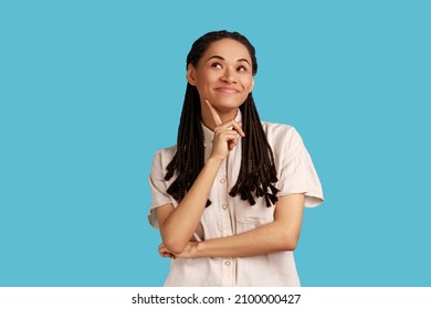 Portrait of pretty woman with dreadlocks, having satisfied expression, keeps finger on chin, concentrated away, thinks about pleasant moment in life. Indoor studio shot isolated on blue background.