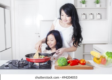 Portrait Of A Pretty Woman And Her Daughter Cooking Vegetable With A Frying In The Kitchen