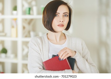 Portrait Of Pretty Woman Holding Closed Book In Modern Office Interior. Knowledge And Information Concept 