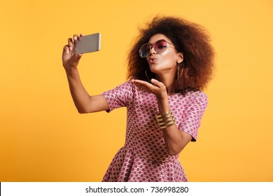 Portrait of a pretty young afro american woman in retro style clothes sending air kiss while standing and taking a selfie isolated over yellow background - Powered by Shutterstock