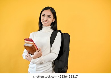 A portrait of a pretty young Asian female college student with her backpack holding her books, standing against a yellow studio isolated background. Education concept - Powered by Shutterstock