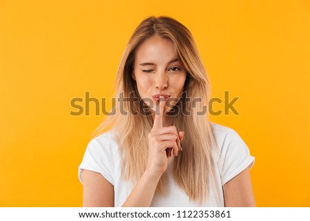 Portrait of a pretty young blonde girl showing silence gesture and winking isolated over yellow background