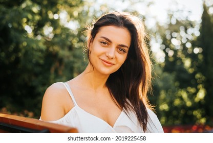 Portrait pretty young brunette woman in white light dress with gentle smile relaxing outdoors and looking at the camera. Beautiful Indian girl with brown big eyes summer lifestyle, close up.
