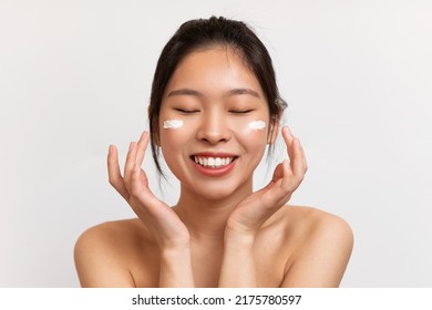 Portrait Of Pretty Young Chinese Lady Applying Moisturizing Cream On Cheeks, Posing Over White Studio Background And Smiling With Closed Eyes