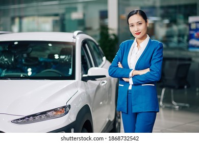 Portrait of pretty young female dealership manager standing next to car, folding arms and looking at camera