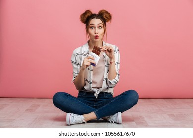 Portrait of a pretty young girl eating chocolate bar while sitting on a floor with legs crossed isolated over pink background