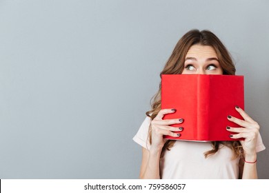 Portrait of a pretty young girl hiding behind an open book and looking away isolated over gray wall background