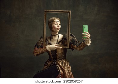 Portrait of pretty young girl, royal person in vintage dress holding picture frame and taking selfie with phone against dark green background. Concept of history, renaissance art, comparison of eras