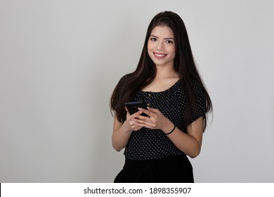 Portrait Of Pretty Young Indian Girl Standing Against White Background And Holding Phone, Smiling And Looking Into Camera.