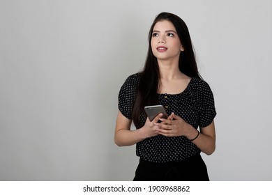 Portrait Of Pretty Young Indian Girl Standing Against White Background And Holding Phone, Smiling And Looking Away From Camera.