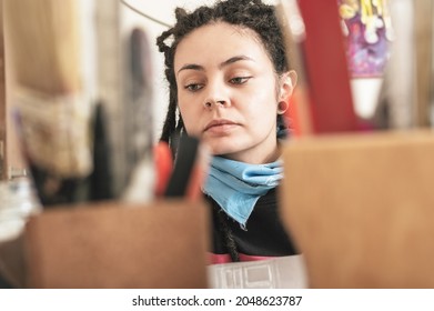 Portrait Of Pretty Young White Hispanic Latin Art Director Girl With Dreadlocks, In Her Work Shop, Looking In Front Of Her For Materials On Her Table To Work On An Art Project.