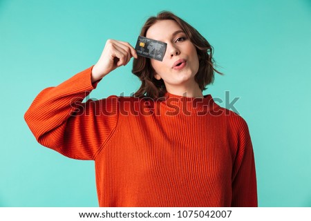 Portrait of a pretty young woman dressed in sweater holding credit card at her face isolated over blue background