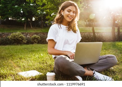 Portrait of pretty young woman sitting on green grass in park with legs crossed during summer day while using laptop and wireless earphone for video call