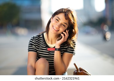 Portrait Of Pretty Young Woman Sitting Outside Talking On Mobile Phone And Looking At Camera