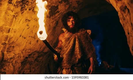 Portrait of Primeval Caveman Wearing Animal Skin Exploring Cave At Night, Holding Torch with Fire Looking into Camera at Night.