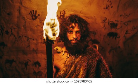 Portrait of Primeval Caveman Wearing Animal Skin Standing in His Cave At Night, Holding Torch with Fire. Primitive Neanderthal Hunter / Homo Sapiens At Night Alone. In the Background Cave Art Drawings