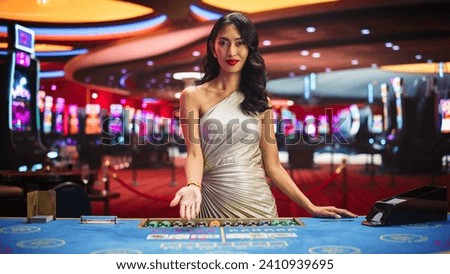 Portrait of a Profesional Asian Croupier in a Casino Dealing Playing Cards on a Baccarat Table. Game Dealer Opens and Reveals Winning Cards to the Audicence, Looking at the Camera in Online Casino