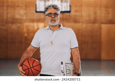 Portrait of a professional basketball trainer on basketball court holding a clipboard and a ball while looking at the camera. A basketball coach ready for coaching basketball players on court. - Powered by Shutterstock