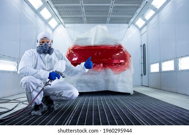 Portrait of professional car painter with protective clothing and mask holding thumbs up and standing by automobile in the painting chamber.