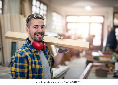 Portrait of professional carpenter holding wood plank material on his shoulder and smiling. Carpentry woodworking workshop in background.