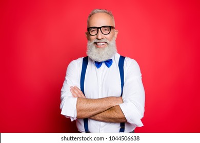 Portrait of professional, cool, old man with beaming smile having his arms crossed, looking at camera, wearing blue bow-tie and suspenders, isolated on red background - Powered by Shutterstock