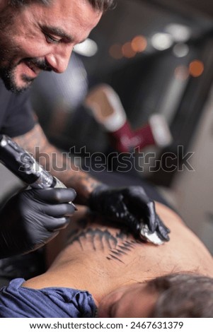 Portrait of a professional creative tattoo master in gloves doing wing tattoo on the back of a young woman in a tattoo parlor. Process of making tattoos with a machine.