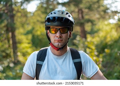 Portrait Of A Professional Cyclist In A Helmet And Glasses With A Serious Face In The Mountain Forest.