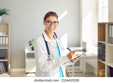 Portrait Of Professional Doctor At Work. Happy Woman In Glasses And Lab Coat Uniform With Stethoscope Standing In Clinic Staff Room Interior, Taking Notes On Clipboard, Looking At Camera And Smiling