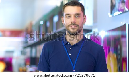 Portrait of a Professional Expert Consultant Smiles and Looks into Camera as Stands in the Bright, Modern Electronics Store Full of Latest Models of TV Sets, Cameras, Tablets and other Devices.