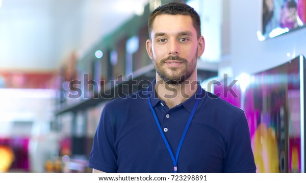 Portrait of a Professional Expert Consultant\
Smiles and Looks into Camera as Stands in the Bright, Modern\
Electronics Store Full of Latest Models of TV Sets, Cameras,\
Tablets and other\
Devices.