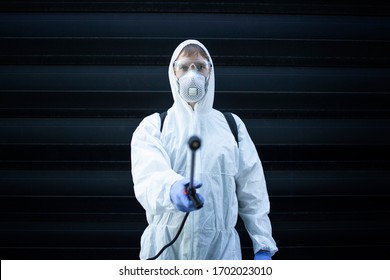 Portrait Of Professional Exterminator Pointing Sprayer Nozzle For Pest Control To The Camera. Man In White Protective Suit Spraying Chemicals.