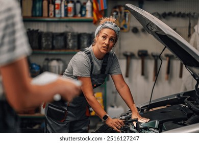 Portrait of professional female mechanic workshop worker standing at auto repair shop with lamp in hand near opened car hood and doing car engine diagnostics with colleague with tablet in foreground. - Powered by Shutterstock