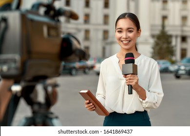 Retrato de una reportera profesional en el trabajo. Joven asiática parada en la calle con un micrófono en la mano y sonriendo a la cámara. Toma horizontal. Atención selectiva a la mujer
