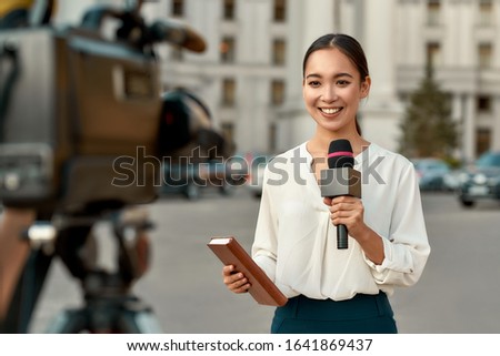 Portrait of professional female reporter at work. Young asian woman standing on the street with a microphone in hand and smiling at camera. Horizontal shot. Selective focus on woman