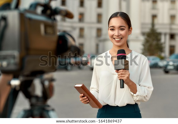 Portrait of\
professional female reporter at work. Young asian woman standing on\
the street with a microphone in hand and smiling at camera.\
Horizontal shot. Selective focus on\
woman