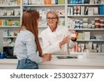 Portrait of professional female senior pharmacy store worker standing behind counter at apothecary and selling beauty product to young woman. Friendly pharmacist helping woman with anti age creme.