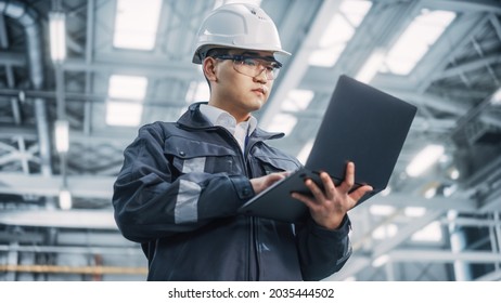 Portrait Of A Professional Heavy Industry Asian Engineer Worker Wearing Safety Uniform And Hard Hat Uses Laptop Computer. Confident Chinese Industrial Specialist Standing In A Factory Facility.