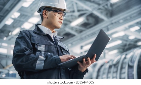 Portrait Of A Professional Heavy Industry Asian Engineer Worker Wearing Safety Uniform And Hard Hat Uses Laptop Computer. Confident Chinese Industrial Specialist Standing In A Factory Facility.