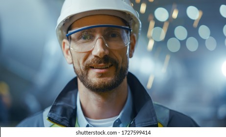 Portrait of Professional Heavy Industry Engineer / Worker Wearing Safety Uniform, Goggles and Hard Hat Smiling. In the Background Unfocused Large Industrial Factory where Welding Sparks Flying - Shutterstock ID 1727599414