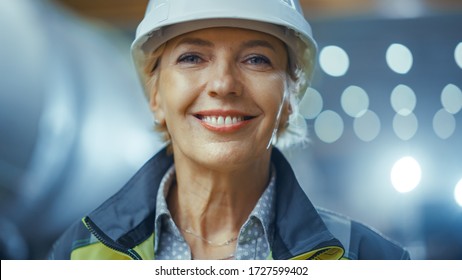 Portrait of Professional Heavy Industry Female Engineer Wearing Safety Uniform and Hard Hat, Smiling Charmingly. In the Background Unfocused Large Industrial Factory where Welding Sparks Flying - Powered by Shutterstock