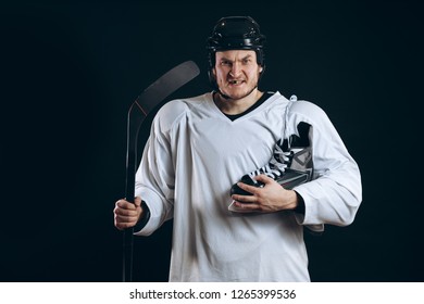 Portrait Of A Professional Hockey Player In A Protective Sportswear And Helmet Posing With Hockey Stick And One Skate In Hands On A Black Background. Sportsman Grins Demonstrating Missing Tooth.