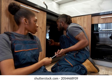 Portrait Of Professional Male African American Plumber With Young Female Apprentice Sitting On Floor In Kitchen. Handyman Checking Pipe Under Sink Using Flashlight, Helper Passing Him Spanner.