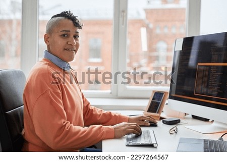 Portrait of professional programmer smiling at camera while writing codes on computer sitting at his workplace