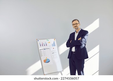 Portrait of professional sales or marketing manager in front of office whiteboard. Happy confident mature business man in suit and glasses standing by office board on light gray wall background