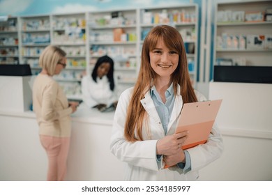 Portrait of professional smiling apothecary worker in lab coat standing at pharmacy shop with clipboard in hands and smiling at camera. In a blurry background are diverse pharmacist and senior patient - Powered by Shutterstock