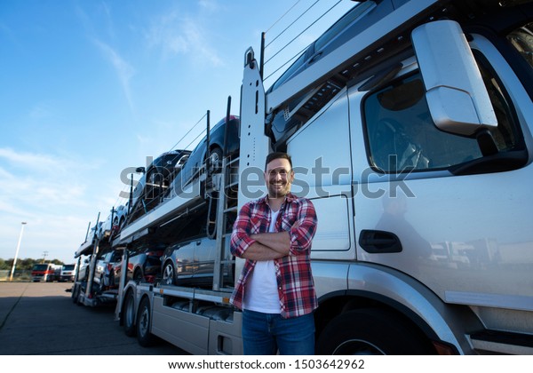 Portrait of professional smiling truck driver with\
crossed arms transporting cars to the market. In background truck\
trailer with cars.