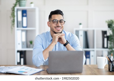 Portrait Of Professional Young Arab Guy Sitting At His Desk With Laptop Pc In Modern Office. Millennial Businessman Using Computer For Online Job At Workplace, Having Virtual Meeting