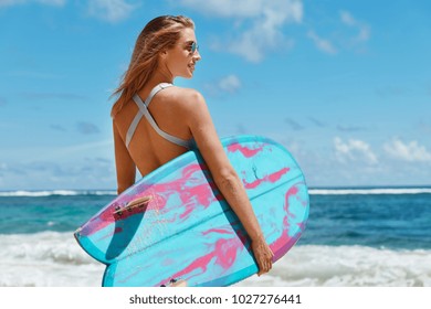 Portrait of professional young female surfer in sunglasses, holds blue big surf board, stands near beautiful ocean view, admires wonderful sunny summer weather, dreams about something pleasant
