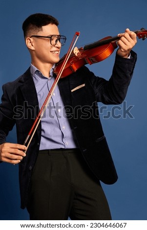 Portrait with professional, young musicians, violinist wearing costume holding fiddlestick and playing on violin and smiling over blue background. Concept of music, art, hobby, human emotions, ad