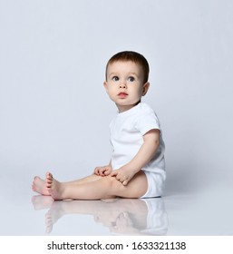 Portrait In Profile Of Calm Infant Baby Boy Toddler In White Bodysuit Sitting Sideways On The Floor Looking At Us On A Gray Background